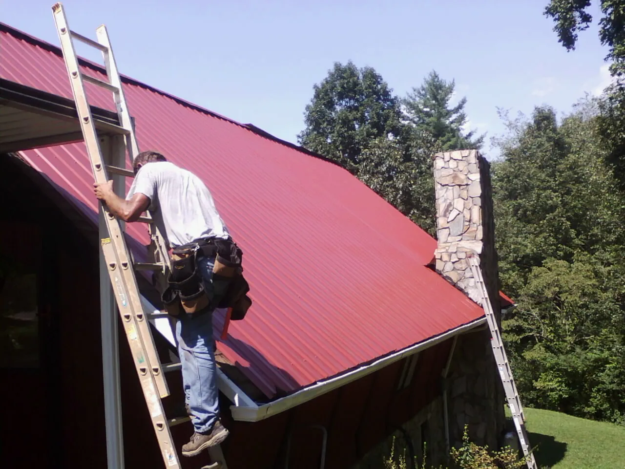 Roofer on a ladder beside a red metal roof and stone chimney — Soffit & Fascia Repair by Roofing Brothers in Whitinsville
