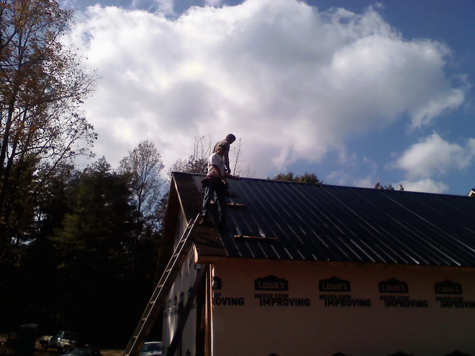 Roofer fastening dark metal panels high against a cloudy sky, from a Roof Inspection job in Whitinsville